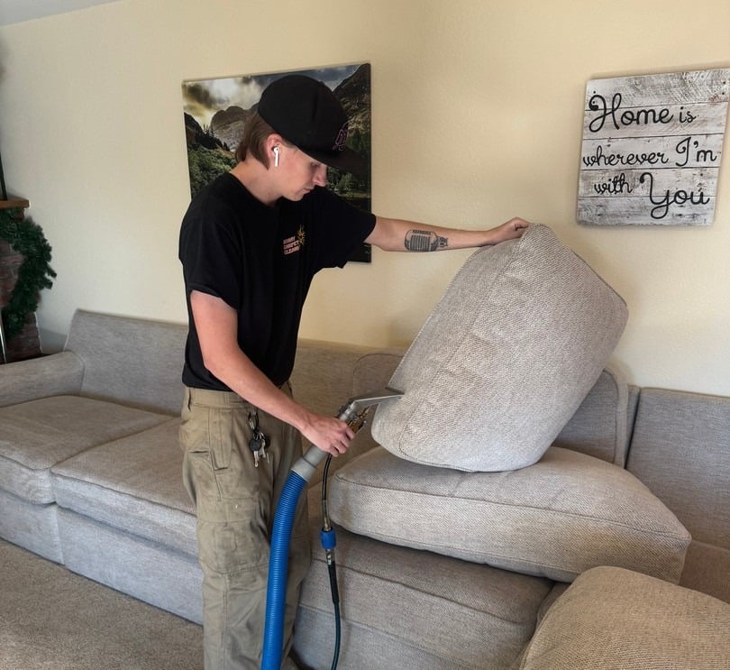 Professional technician using upholstery cleaning equipment on a light-colored sofa cushion in a cozy living room setting.