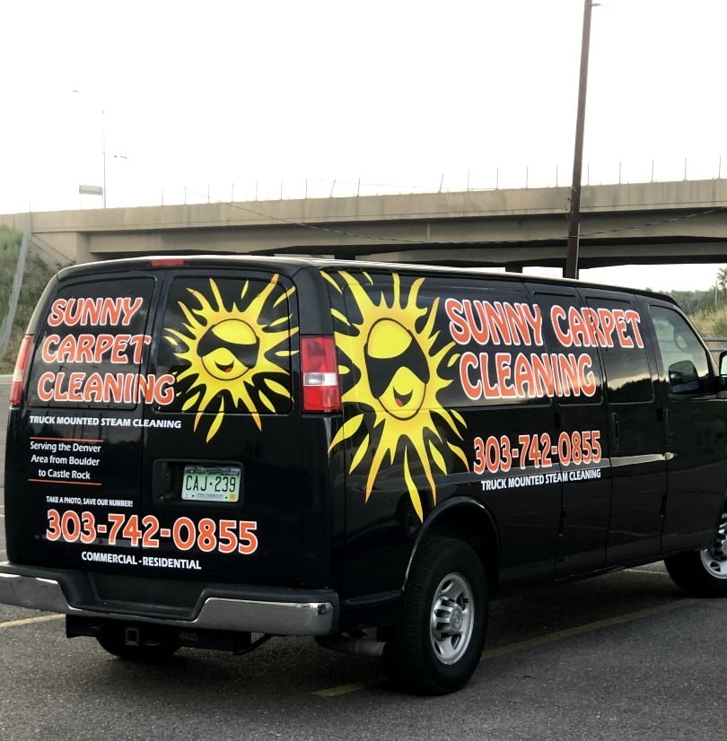 IMG_0996 Sunny Carpet Cleaning service van with bright yellow sun logo, contact number, and text promoting truck-mounted steam cleaning, parked under a highway overpass.